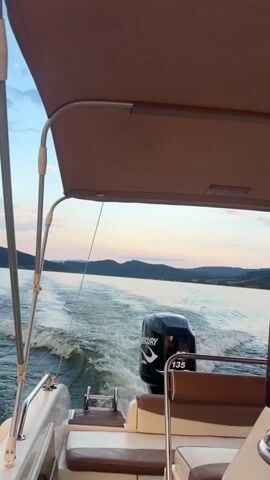 The video captures a rear view of a boat moving across a body of water. The boat has a canopy overhead and brown upholstered seating. A black Mercury outboard motor with the number 135 is visible at the stern of the boat, churning up a wake in the water. In the distance, hills or mountains line the horizon under a sky with scattered clouds, suggesting either sunrise or sunset. The water appears calm except for the wake. No people are visible in the frame.
