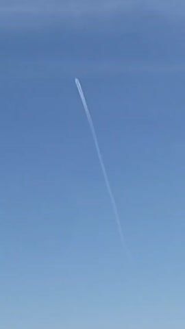 A contrail stretches upward through a clear blue sky, bisecting the frame. An airplane is at the leading point of the contrail, in the upper portion of the view.