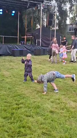 The video shows a group of people on a grassy area in front of a stage. Two young boys are in the foreground, with one boy in a camouflage jacket and denim shorts and the other in a patterned tracksuit. The boy in the tracksuit is doing a handstand, while the boy in the camouflage jacket is running. In the background, several adults and children are dancing. The stage is set up with speakers and lighting equipment. The scene appears to be an outdoor event, possibly a concert or festival.
