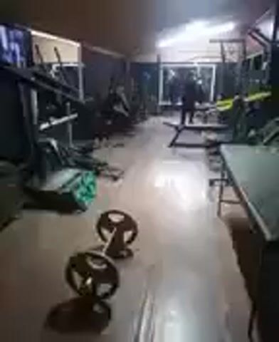 The video shows a dimly lit gym with various weightlifting equipment. In the foreground, a barbell with weight plates is visible on the floor. The background reveals rows of exercise machines, benches, and mirrors. The lighting appears to be artificial, possibly from fluorescent tubes on the ceiling, casting shadows across the room. The overall impression is of a functional, albeit possibly older or less well-maintained, gym.