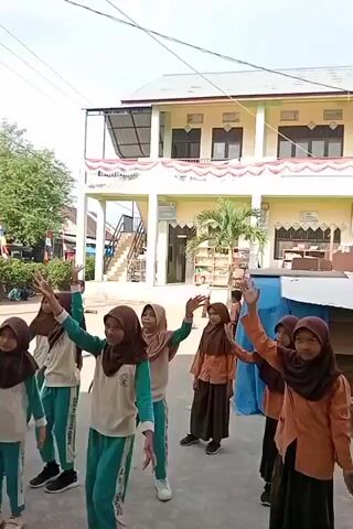 A group of young girls, dressed in school uniforms with hijabs, are engaged in a dance activity outdoors. They are arranged in a semi-circle facing a building, with some in the foreground and others slightly further back. The building appears to be a two-story structure with a beige facade, white trim, and red and white bunting decorating the upper section. Some windows are visible on the upper floor. The ground is paved, and there are trees and other structures in the background, suggesting a schoolyard or open area. The girls are performing synchronized movements with their arms raised and bodies in motion. The lighting suggests it is daytime, with sunlight casting shadows.