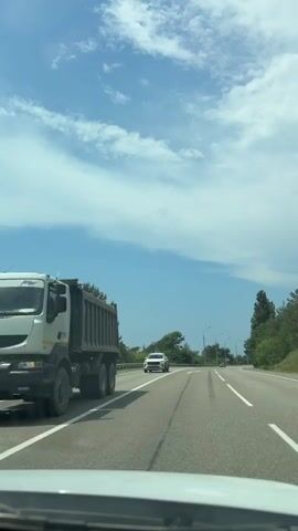 The video shows a view from inside a vehicle driving on a highway. The highway is paved with asphalt and has white lane markings. There are trees and greenery on both sides of the road, and the sky is blue with scattered clouds. Other vehicles are visible on the road, including a white SUV and a black car. The video appears to be filmed on a sunny day.