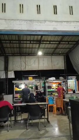 The video captures an outdoor food stall scene at night. The stall area is covered by a metal roof, and underneath are several food carts where people are working. Two individuals are seen preparing food at the carts, with one wearing a red shirt with the number 6 on the back. Customers are seated at tables, including one person in a red shirt and another wearing a hijab. A parked motorcycle is also visible in the area. The building’s facade is white with rectangular openings near the roofline.