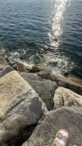 A first-person perspective video shows a rocky shoreline and the ocean. The camera angle is angled down towards the water, with large grey and tan rocks in the foreground and a small portion of a sandal visible at the bottom right corner. The ocean is dark blue with sunlight reflecting off the surface, creating a shimmering effect. Small waves are crashing against the rocks. In the distance, the horizon shows a hazy line of mountains or landmasses under a bright blue sky with a few scattered clouds. The sun is visible in the upper left portion of the frame, casting a bright glare on the water. The overall scene depicts a calm and sunny day at the seaside.