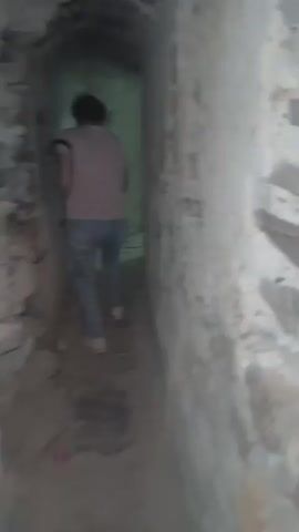 A woman walks away from the camera down a narrow, dimly lit passage. The passage has rough, aged stone walls on both sides and a brick floor. The woman is wearing a sleeveless pink shirt, blue jeans, and white shoes. She is walking away from the viewer, further down the passage, which appears to be part of an old structure like a castle or dungeon. The lighting is poor, creating shadows and obscuring details of the passageway.