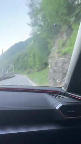 The video shows a view from inside a car driving on a road next to a train track. The car's dashboard, steering wheel, and digital display are visible. The display shows a map and information about the car's speed. Outside the car, there are mountains, a lake, and trees. The driver's hands are visible on the steering wheel. The sky is clear and the sun is setting.