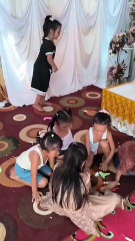 A group of young children are gathered on a patterned carpet. One child stands and faces away from the camera. Five children are seated on the carpet, some are interacting with small objects. A small boy in a red patterned shirt and blue jeans stands in the center of the frame. In the background, a white table with gold trim is covered with a white cloth. There are floral arrangements and white drapery.