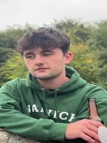 Two young men are standing outside. The man in the foreground is wearing a green hooded sweatshirt with white lettering and is holding a brown beer bottle. The other man wears a dark blue sweatshirt and a light gray baseball cap. The background is a tree line. The man in the foreground says “Fair. Then you gotta ask him have you been shagg-”. The man in the baseball cap laughs.