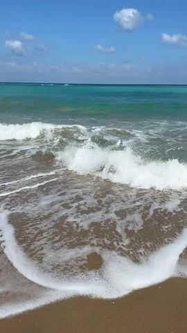 The video captures a serene beach scene at Palmachim, Israel. The camera focuses on the gentle ebb and flow of the ocean waves meeting the sandy shore under a clear blue sky dotted with a few fluffy white clouds. The water exhibits a beautiful turquoise hue, transitioning to a deeper blue towards the horizon. Foamy white surf rushes onto the wet sand, leaving behind intricate patterns as it recedes.

The time of day appears to be midday, indicated by the bright sunlight and the clear, vibrant colors of the sky and sea. The weather seems pleasant and warm, perfect for a day at the beach. There are no people or man-made objects visible in the frame, emphasizing the natural beauty and tranquility of the location. The overall mood evoked is one of peace and relaxation, inviting viewers to immerse themselves in the calming rhythm of the waves. No text is visible in the video.