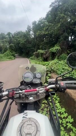 The video pans from the handlebars of a motorcycle parked on the side of a paved road. The motorcycle has a windshield and mirrors visible. The road curves to the left and disappears into a lush green landscape dominated by trees and dense foliage. The environment appears tropical, with tall trees, some resembling palm trees, and thick undergrowth covering the ground and climbing on structures. In the background, beyond the trees, a bright, overcast sky is visible. A partially constructed structure, possibly a shelter, made of wood and covered with purple tarps, is visible nestled among the greenery. The ground is a mix of dirt and red soil. The overall impression is a natural, somewhat wild setting, possibly a rural road in a tropical region.