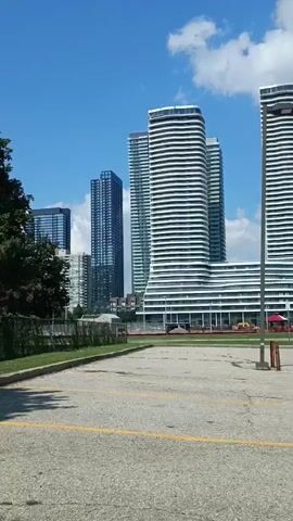 This is a panning video shot from a parking lot. In the background, a city skyline is visible with several modern, high-rise buildings. Some of these buildings have distinctive wavy or curved facades, particularly two prominent white towers in the center. The sky is blue with scattered white clouds. The parking lot in the foreground is paved with asphalt and marked with yellow lines. There is some overgrown grass and a fence to the left of the frame. A single lamppost stands in the middle of the parking lot. The overall impression is a clear, sunny day in an urban or suburban environment.
