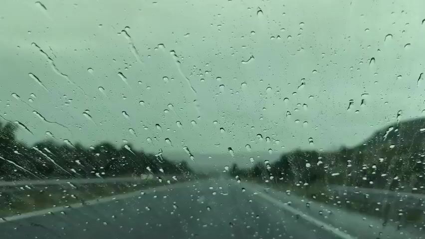The video shows a view through a car windshield during rainfall. The windshield is covered in raindrops, some of which are streaking down the glass. The road ahead is visible, with trees lining either side. The sky is overcast and appears to be a light grey color. The overall impression is one of driving in inclement weather.