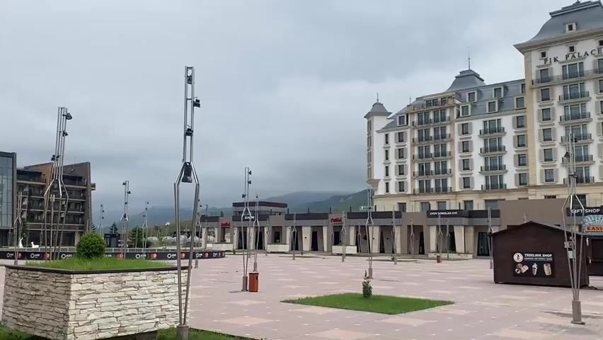 The video shows an outdoor plaza in front of a large building labeled "FIK PALACE." The plaza is paved with square tiles in a reddish-brown color. There are several small, wooden kiosks or stands in the plaza. One kiosk has a sign with an image of ice cream and text that is partially visible. Another kiosk has a sign that says "Kartoff". There are a few small green grassy areas with young trees planted in them. Several tall, slender lamp posts are scattered throughout the plaza. The building in the background has many windows and balconies, and appears to be a hotel or resort. In the distance, there are mountains, and the sky is overcast. There are no people visible in the video.