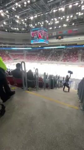The video shows a large indoor ice hockey arena with rows of red seats largely filled with spectators. A central screen displays video footage. A person dressed in black with pom-poms is standing between the railing and seats, seemingly performing or cheering. Another person is standing nearby wearing a neon green jacket. People sit watching the event. A stroller is visible on the left. Overhead, bright lights illuminate the arena.