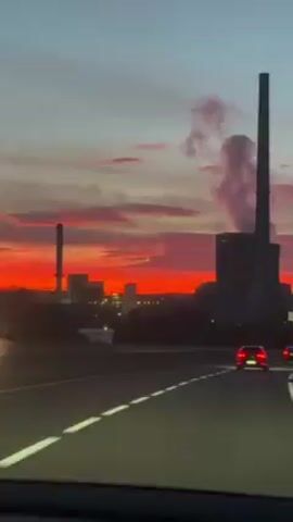 The video shows a view of a city skyline at sunset from what appears to be a moving car. The sky is a mix of red, orange, and gray, with clouds stretched across the horizon. The city skyline is silhouetted against the bright sky, with several buildings and a tall smokestack visible. The car is on a road with white lane markers. The video is shaky, likely due to the movement of the car. There is an audible voice speaking in French.