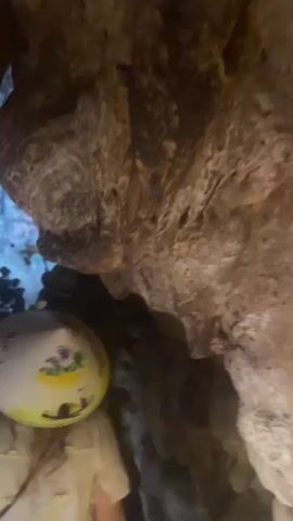 The video shows the interior of a cave with illuminated rock formations and stalactites. A person wearing a conical hat is in the foreground. In the background, other people are visible, some standing and some walking, with lights illuminating the path. The cave walls are rough and textured, with varying shades of brown and gray. Some areas of the cave appear wet, reflecting the light. The overall impression is of a tourist attraction within a natural cave system.