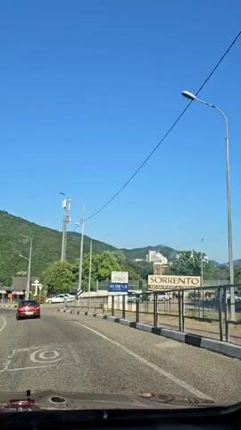A video captures a drive through a sunlit area with a clear blue sky. The camera appears to be mounted inside a vehicle, looking forward and slightly to the right. The road ahead curves gently to the left.

On the right side of the road, there's a black and white striped barrier, followed by a fence. Beyond the fence, a large sign with yellow lettering reads "SORRENTO RESTAURANT & BAR." Further to the right, a few white cars are parked. In the distance, buildings are visible on a hillside, suggesting a populated area.

On the left side of the road, a red car is driving away from the camera. Further down, there's a tall cell phone tower. Next to it stands a large white cross, indicating a religious site. Trees and greenery are abundant, with a lush, green hillside rising in the background. The sun is high in the sky, suggesting it's midday or afternoon, and the weather appears to be clear and warm. The overall mood is peaceful and scenic, typical of a drive through a coastal or mountainous region. There's also another sign partially visible with the word "HIDENS" on it.