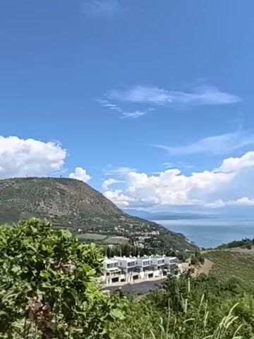 The video pans from left to right across a scenic landscape featuring a body of water, a coastline with hills, and a few modern buildings. In the foreground, there are trees and tall grasses. The sky is blue with scattered white clouds. The water appears calm, reflecting the blue of the sky. The coastline is dotted with greenery, and a small cluster of white, multi-story buildings is visible on a hillside overlooking the water. Further in the distance, more hills and a hazy horizon are discernible. The overall impression is one of a peaceful, sunny day in a coastal region.