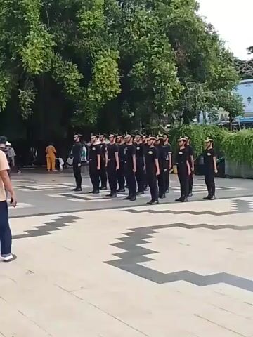 Here's a description of the video:

A group of about 15-20 people, seemingly dressed in black uniforms with hats, are standing in formation on a paved area. The ground has a geometric pattern made of light and dark stones. The group is standing at attention. In the background, there are trees and people walking around. A person dressed in civilian clothes (blue jeans, white shoes) can be seen in the lower left of the frame, standing slightly apart from the group.  There's also a billboard in the background and some plants. The overall setting appears to be a public square or park area.