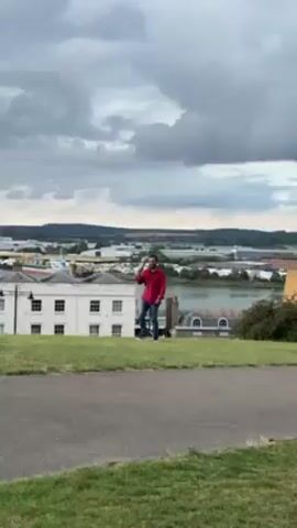 A man in a red shirt and jeans is walking on a grassy hill overlooking a town. He is holding a phone or similar device in his hand. The sky is overcast with dark clouds. The town below includes white buildings with black windows and a body of water with a port and industrial buildings in the background. The man is walking towards the right side of the frame. A tree is visible in the foreground.