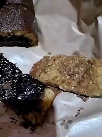 This media file appears to be a close-up video of a dessert, likely Martabak Manis, a popular Indonesian sweet pancake. The camera focuses on several cut pieces of the dessert, arranged on white paper. One piece is a thick, folded slice with a dark, granular filling, possibly chocolate or nuts, visible between layers of the cooked pancake. Another piece is lighter in color, with a slightly textured surface and some scattered toppings, perhaps sugar or cheese. A third piece is darker and appears to be a different variety, possibly a chocolate-filled version. The lighting is dim, suggesting an indoor setting, and the focus is entirely on the food. There are no people visible, and no discernible text in the video. The video appears to be a brief, casual recording of a food item, possibly intended to showcase its appearance or for personal documentation. The context of Palangka Raya, Indonesia, aligns with Martabak Manis being a local delicacy.