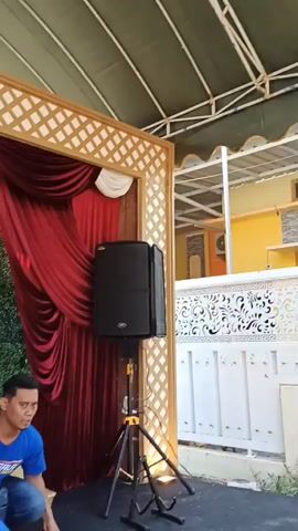 The video begins with a shot of a black speaker on a stand, with red curtains and a trellis in the background. A man wearing a blue shirt and dark pants is visible in the lower left corner, kneeling on the floor. As the video progresses, the camera pans to reveal a large banner that reads "JALAN SANTAI DALAM RANGKA HUT KEMERDEKAAN RI KE-81." Below the banner are several wrapped gifts in various colors, including pink, blue, yellow, and green, as well as a golden gift box. A large blue duffel bag is also visible in the foreground. The camera then pans back to show the speaker and the red curtains again, followed by another view of the banner and gifts. The final shot shows the speaker and the trellis from a slightly different angle. The environment appears to be an outdoor event, possibly a celebration or festival, with the decorations and gifts suggesting a festive occasion.