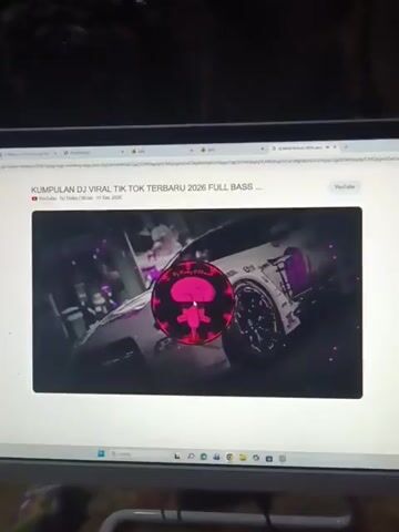 The video shows a laptop screen displaying a YouTube video. The YouTube video features a thumbnail with a graphic of SpongeBob SquarePants' character Squidward Tentacles, surrounded by pulsating colorful lights and what appears to be a car. The title of the YouTube video is "KUMPULAN DJ VIRAL TIK TOK TERBARU 2026 FULL BASS". The surrounding YouTube interface elements like the play button, progress bar, and channel information are also visible. The lighting suggests the room is dimly lit, with the laptop screen being the primary light source.