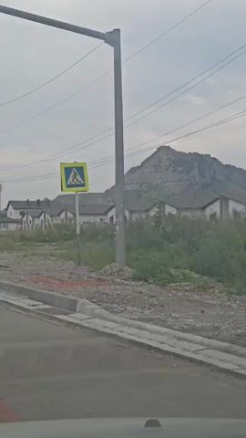 The video pans across a suburban landscape with houses and a mountain in the background. A pedestrian crossing sign is visible on the left, followed by a bus stop sign. The sky is overcast.
