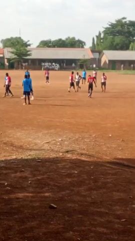 This media file depicts a group of young people playing soccer on a dirt field. The scene appears to be set in Kakuri Gwari, Nigeria. The time of day seems to be late afternoon, judging by the long shadows cast on the field and the soft lighting. The sky is partly cloudy, suggesting fair weather.

The subjects are primarily young individuals, most of whom are wearing athletic attire, specifically soccer uniforms in shades of blue and red. There are approximately 10-15 individuals visible on the field, actively engaged in a soccer game. One person in a blue uniform is bending over, possibly attending to an injured player or retrieving the ball. Another person in a blue uniform is holding a soccer ball. Several other players are positioned around the field, some running and others standing. In the background, there are school buildings with corrugated iron roofs and some trees. A group of people are gathered in front of one of the buildings, possibly spectators.

The activity is clearly a soccer match or practice session. The participants are exhibiting a range of postures and actions consistent with playing the sport, including running, kicking, and interacting with the ball. There is no visible text in the media file. The overall mood of the scene appears to be active and energetic, with a sense of friendly competition or organized sport. The dirt field, simple buildings, and clear sky are characteristic of many outdoor community settings in the region.