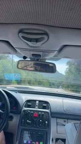 This media file is a first-person view from inside a car, showing the dashboard, windshield, and the road ahead. The driver, a person wearing glasses, is visible in the rearview mirror. The car is driving on a paved road that winds through a lush, green landscape with trees and hills. The sky appears clear and blue, suggesting a sunny day.

The dashboard of the car is visible, with a steering wheel on the left and a central console featuring climate controls and a modern infotainment system. The infotainment screen displays the time as "12:14" and the date as "July 27, 2020." A hazard light button is prominently displayed.

Outside the car, on the left side of the road, a blue sign with white symbols is visible, one of which resembles a wheelchair symbol, indicating accessibility features. The road ahead stretches into the distance, with trees lining both sides. The overall scene suggests a pleasant drive through a scenic rural or coastal area, likely in Italy given the location context of Punta Ala. The sunlight and clear sky indicate good weather. The mood conveyed is relaxed and perhaps leisurely, typical of a road trip or a scenic drive.
