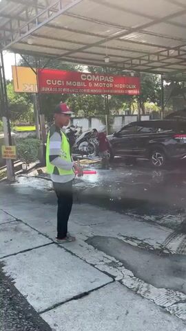 The video shows a car wash facility. A person wearing a red hat and a green vest is holding a hose and spraying water. Several cars and motorcycles are present, some being washed. The facility has a roof structure with a sign that reads "COMPASS CUCI MOBIL & MOTOR HIDROLIK". There are other people in the background, likely workers at the car wash. The ground is wet from the water, and there are puddles on the concrete. The environment appears to be outdoors with trees and vegetation visible in the background.