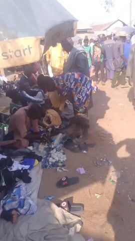 The video shows a bustling outdoor market scene in what appears to be a West African country.  Several vendors are displaying goods, mainly clothing and shoes, on blankets or tarps spread on the ground. People, dressed in a variety of colorful traditional and modern clothes, are milling about, browsing and possibly purchasing items. There are open umbrellas providing shade. One vendor sits on the ground, sorting through items. A blue and white Momo Payment Service Bank umbrella is visible in the background. The ground appears to be dusty and unpaved. Clothes are also displayed on racks.  There is a water tower in the background.