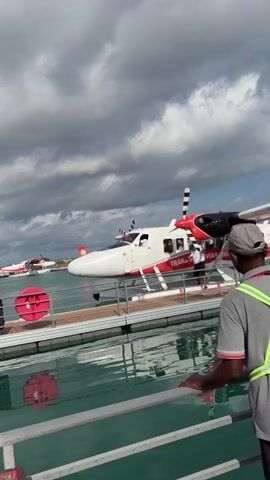 A group of people are gathered on a dock next to a seaplane. The seaplane is white with red accents and the words "Trans Maldivian Airways" printed on it. There are two seaplanes visible in the background. The sky is cloudy and the water is a clear turquoise. A man in a red hat appears to be directing the passengers. One of the passengers is holding a stuffed animal. The dock is made of wood and has metal railings.