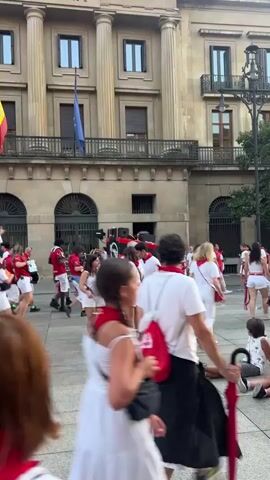 The video shows a crowd of people in a plaza, in front of a large building with classical architecture. Most of the people are wearing white shirts and shorts, and many have red scarves around their necks. Some individuals are playing musical instruments, such as trumpets. There is a small cart or vehicle with speakers on it in the background. People appear to be walking, talking, and interacting with each other. The overall atmosphere seems festive, possibly a parade or a celebration. Flags are visible on the building, including the European Union flag and the Spanish flag. The ground is paved.
