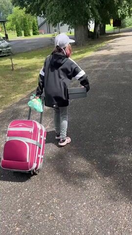 A person wearing a baseball cap, black hoodie with white accents, grey pants, and pink crocs walks away from the camera pulling a maroon suitcase on wheels. They are also carrying a white plastic bag in their left hand and a black box in their right hand. The person is walking on an asphalt path, which appears to be a road or driveway. To the left of the path is a grassy area with trees. In the background, there are houses and more trees. The weather appears to be sunny and warm.