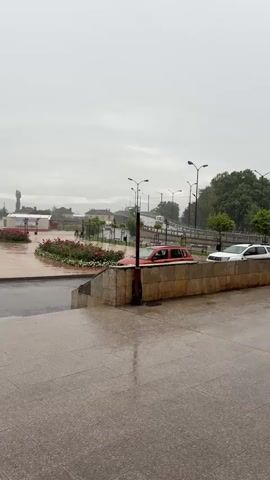 The video displays an overcast daytime scene of a town square or park in Gara Elin Pelin, Bulgaria, characterized by wet surfaces suggesting recent or ongoing rain.

In the immediate foreground, there is a wet, light-colored tiled pavement. A low stone wall separates this area from a paved road where a red SUV is partially visible, parked facing left, and a white car is seen further back.

The midground reveals a spacious public square featuring organized flower beds filled with pink and white roses, interspersed with small, manicured trees and a series of black streetlights. An active fountain is visible in the distance within the square.

As the camera pans left, a white church building with red domes and a prominent cross on its main dome becomes visible, surrounded by lush green trees and shrubs. Several vehicles, including a blue car and a white van, are parked near the church. The background consists of various buildings, a bridge, and hazy distant hills or mountains. A tall cellular tower stands on the far left.

No individuals are visible in the scene, and no specific events or activities are taking place, contributing to a quiet and still atmosphere under the gray sky. There is no discernible text visible in the video.