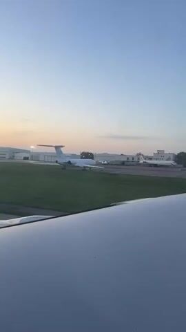 The view from inside an airplane reveals the wing of the aircraft and a landscape containing two other airplanes parked on the runway, set against a backdrop of buildings and grass. The scene is lit by an early morning or late evening light, suggesting dawn or dusk. The sky above is a gradient of blue and faint orange. The airplanes in the background are white and clearly visible, and the buildings are white with closed bay doors. The scene is serene and peaceful, as the aircraft remains stationary.