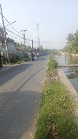 The media file shows a scene in Sarakan, Indonesia. The camera is positioned at a slight angle, looking down a road next to a canal. The time of day appears to be morning or late afternoon, judging by the shadows cast by the trees and utility poles. The weather seems clear and sunny.

The main subjects are people, who are in the canal, seemingly playing or swimming. There are several individuals visible, some appearing to be children. On the road, there are a few motorcycles, suggesting local transportation. The setting is a rural or semi-urban area, with houses and shops lining the side of the road opposite the canal. Utility poles with extensive wiring are also prominent, indicating infrastructure. Lush green vegetation, including palm trees and other tropical plants, borders the canal and the road, adding to the tropical feel of the location.

Notable details include the concrete embankment of the canal and the asphalt road. There are some pieces of trash and debris along the edge of the road near the canal, a common sight in many developing areas. The overall mood of the scene is peaceful and ordinary, depicting a typical day in the community. No visible text is present in the media file.