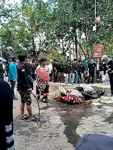 The video shows an outdoor performance or cultural event. A person in traditional costume with a headdress and colorful leg coverings is in the center of the frame, holding red tassels. Another person, dressed in dark clothing, stands to the left, holding a whip. Several other individuals, many wearing black shirts with emblems, are gathered around, observing. In the foreground, two individuals are lying on the ground, one appearing to be partially submerged in water or a dark liquid from a black basin. A bucket is being tossed in the air, splashing water. The environment is outdoors with trees and what appears to be a street or paved area. The overall scene suggests a traditional folk performance that involves water and potentially a theatrical element.