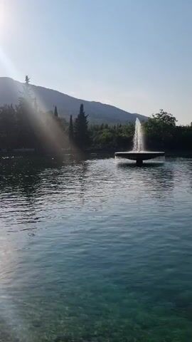 The video shows a fountain in a body of water. The water is clear and the bottom is visible, with rocks and pebbles. In the background, there are trees and mountains. The sky is clear and blue, with the sun shining. There are no people or animals visible in the video.