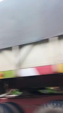 The video appears to be shot from the perspective of someone traveling in a vehicle. The initial frames show a blurry, out-of-focus view of the side of a vehicle, possibly a truck, with a cream-colored cab. Green foliage and palm trees are visible in the background, suggesting a tropical or subtropical environment. The camera then pans and shows a blurry landscape with trees and some structures that appear to be simple buildings, possibly homes, with corrugated metal roofs. The ground is a reddish-brown color, indicating it might be dirt or clay. The sky is overcast with some patches of sunlight breaking through the clouds. The overall impression is of a rural or semi-rural setting, possibly in a developing country, with the motion blur indicating movement.