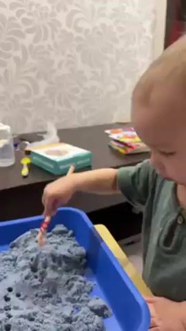 A young, light-skinned child with blond hair is seen in a close-up, playing with kinetic sand in a blue plastic tray on a yellow surface. The child is wearing a green, short-sleeved shirt. The child uses a small wooden stick to interact with the sand. In the background, a dark wooden surface holds various items, including a water bottle, a book or game box, and colorful items that seem to be a children's educational toy. The wallpaper behind is patterned with a swirling, floral design. Another person's hand enters the frame and puts another wooden stick into the blue tray.