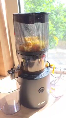 A Hurom brand juicer is situated on a wooden surface by a window. The juicer is black and cylindrical, with a clear plastic top compartment containing a yellowish pulp. A spout extends from the side of the juicer, and a clear plastic container is positioned beneath it, collecting some liquid. The background shows a blurred view of green foliage through the window, suggesting an outdoor environment. The lighting is bright, indicating daytime. No people are visible in the frame.