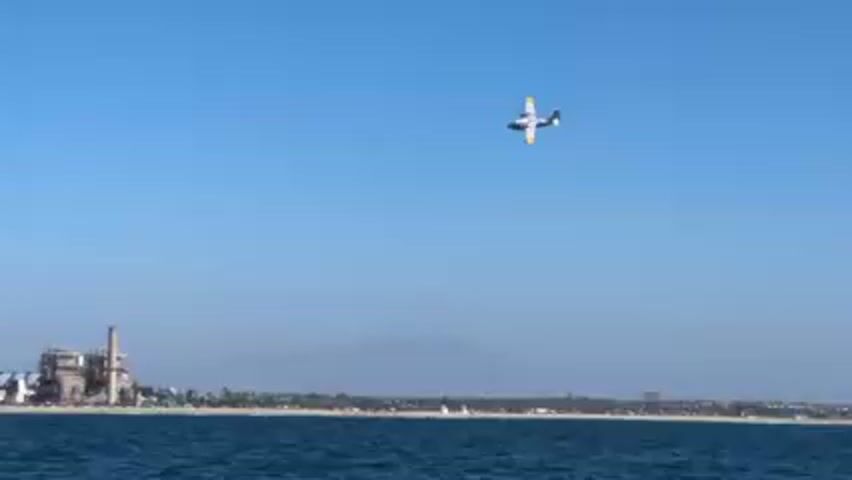 The video shows a body of water with a shoreline in the background. A large industrial complex with smokestacks is visible on the shore. A small, white and yellow airplane flies in the clear blue sky above the shoreline and the water. The water is choppy with small waves. The shoreline appears to be a beach with vegetation and possibly buildings beyond. Mountains are faintly visible in the far distance.