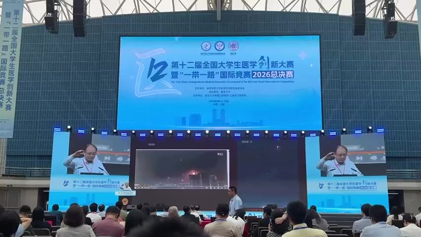 A large screen displays the title "The 12th China Undergraduate Medical Innovation Competition & The Belt and Road International Competition 2026 Final." Below the main screen, two smaller screens show a man speaking at a podium, saluting with his right hand to his forehead. The man appears to be wearing a light-colored shirt and glasses. In the center, a third screen shows a video of what appears to be a fireworks display or a nighttime cityscape with lights. A live audience is seated in front of the stage, facing the screens. Some individuals are standing and walking in the aisles between the audience seats. The environment appears to be a large indoor venue with a high ceiling and visible structural beams. The overall lighting is bright, with spotlights directed at the stage and screens. The lower part of the screens and the stage area have blue lighting elements.