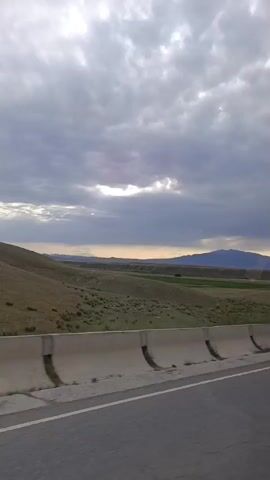 A scenic view of rolling hills and mountains under a cloudy sky, with a concrete barrier and a road in the foreground. The camera is in motion, suggesting it is mounted on a vehicle. The landscape appears to be rural and undeveloped. The lighting is dim, indicating it may be early morning or late evening. There are no people or other vehicles visible in the frame.