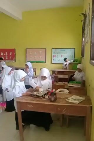 The video shows a classroom setting with several students, mostly wearing white uniforms and headscarves. The classroom walls are painted yellow. There are wooden desks and chairs. In the foreground, two students are working at a desk. One is wearing a white headscarf and appears to be placing items into a tray. Another student beside her is also looking at the tray. In the background, other students are seated at desks, some are writing or looking at papers. Some students are wearing white shirts and dark pants, and a few boys are wearing skullcaps. There are posters and educational materials on the walls. A window is visible on the left side of the frame. The overall atmosphere appears to be one of a classroom activity or lesson.