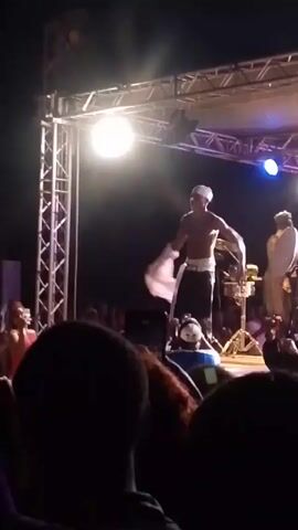 The video shows a performance on a stage at night, likely a concert. The lead performer is shirtless, wearing a white head covering and dark pants. He is holding a white cloth and waving it in the air.  Behind him, a band is visible, some members wearing white shirts. There are stage lights overhead, and a crowd is visible in the foreground, with some people holding up their phones to record the performance.