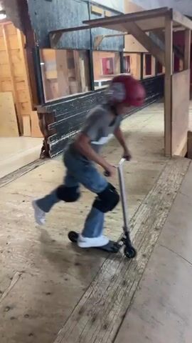 A person wearing a red helmet, gray t-shirt, blue jeans, and knee pads is riding a scooter in a skate park. The person is in motion, with their body angled as they ride down a ramp. The skate park features wooden ramps and other structures. Other people are visible in the background, some sitting on the ramps. The video appears to be a recording of someone performing a scooter trick.
