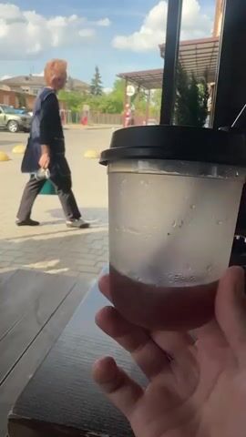 The video shows a close-up of a transparent plastic cup filled with a reddish-brown beverage and ice, held in a person's hand. A black lid with a straw is on top of the cup. The person's thumb is visible. The background is slightly out of focus but shows an outdoor scene.

In the background, a woman with reddish hair wearing a dark vest over a lighter colored shirt and dark pants is walking away from the camera, carrying a blue bucket. She appears to be in a public area, possibly a street or courtyard. Further in the background, there are buildings with brick and light-colored facades, parked cars, and a lamppost. The sky is visible with some clouds. The overall lighting suggests it is daytime, likely late morning or afternoon, with sunny weather. The setting appears to be a town or city in Ukraine, given the location context of Berdychiv. The activity seems to be a casual moment, perhaps enjoying a drink outdoors. There is no visible text in the video.